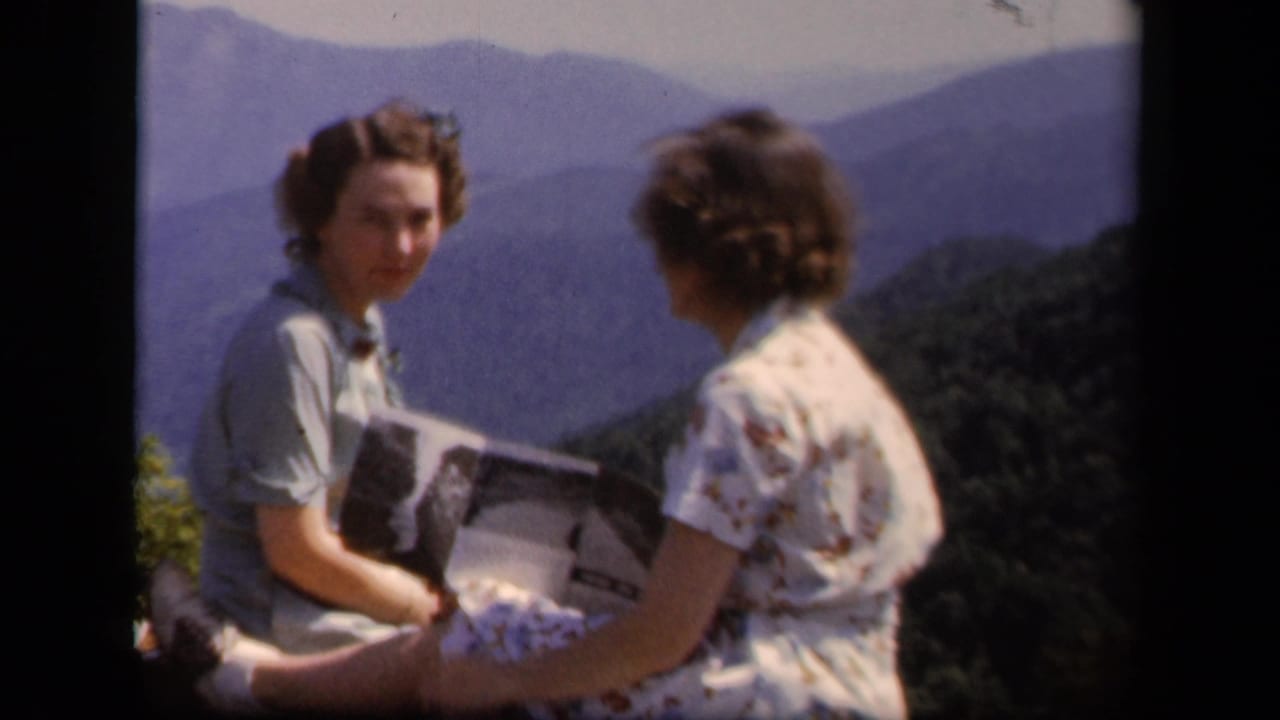 Two women face each other at an overlook, with one holding an open booklet or paper.