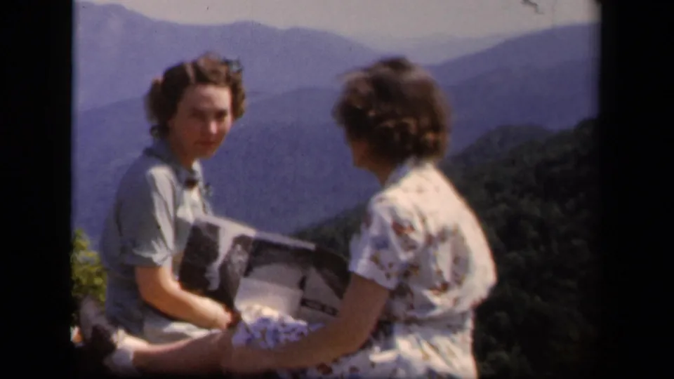 At the Virginia mountain overlook, one woman in a light dress holds an open paper or booklet while another woman in a floral dress sits nearby.