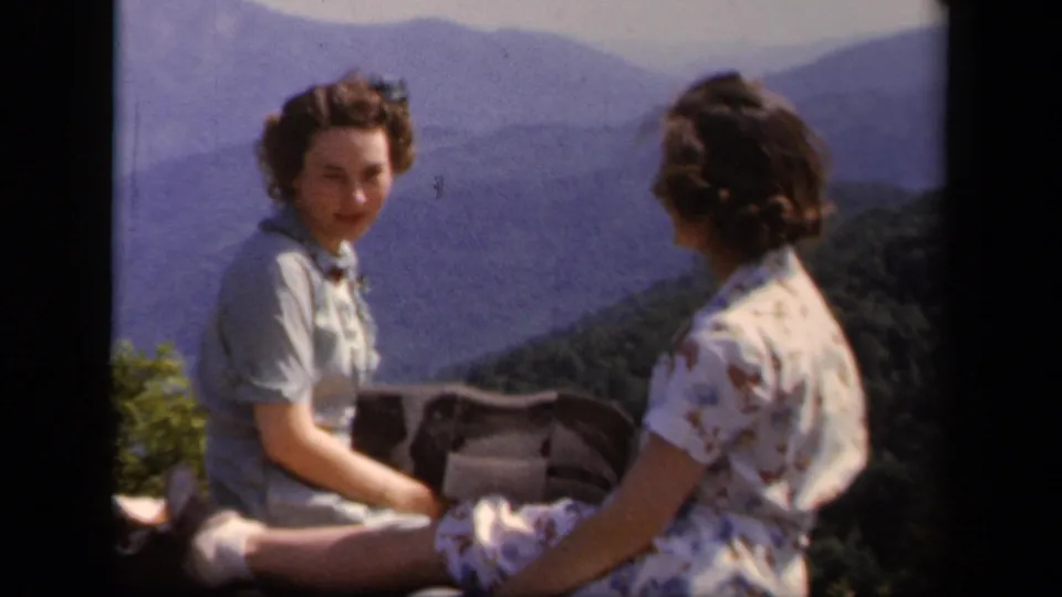 The home-movie frame shows two women seated at Skyline Drive, one facing the camera as the other turns toward her with mountains behind them.