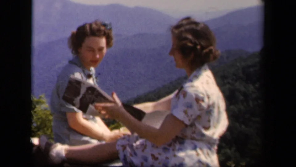 At Skyline Drive in 1943, a woman in a floral dress gestures while speaking with a woman holding a paper or booklet near the overlook edge.