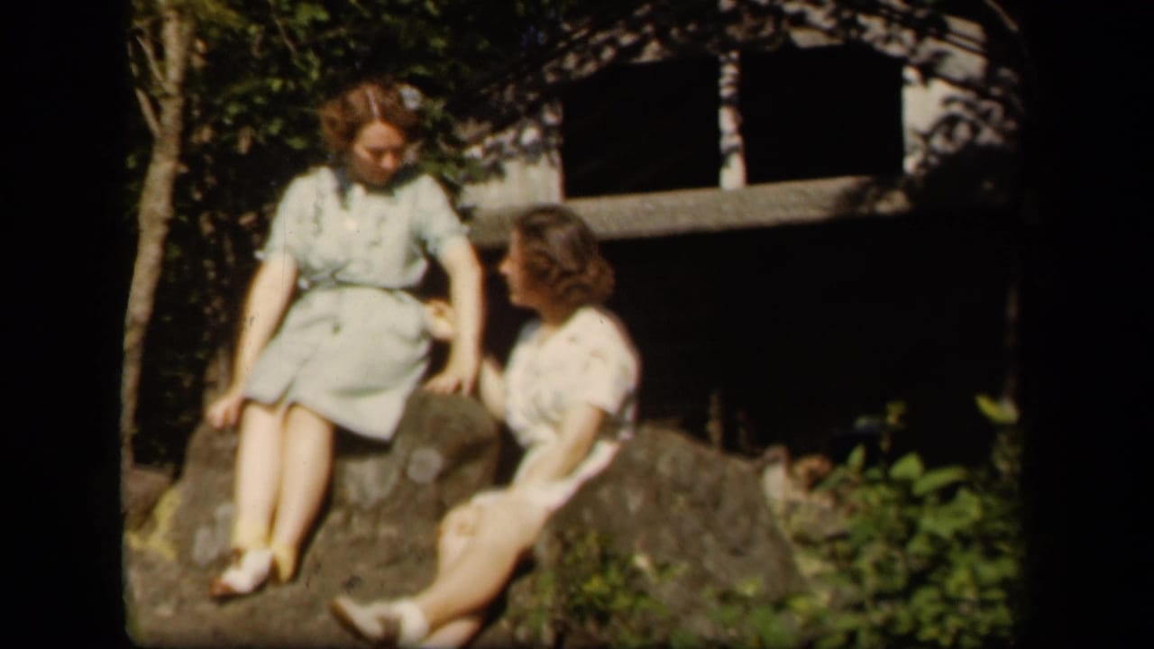 Two women in light dresses sit on rocks outdoors near trees and a dark structure.