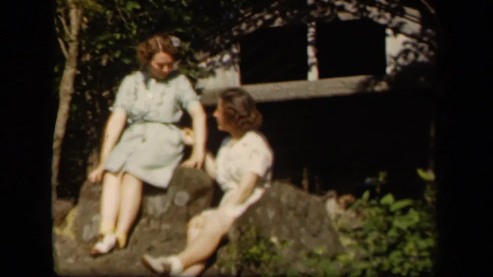 Two young women in 1940s dresses sit on rocks at Skyline Drive, Virginia, with foliage and a shaded structure behind them.