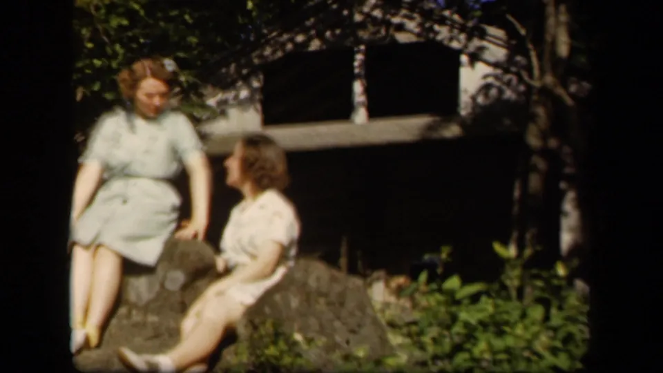 Two young women in period dresses sit on rocks at Skyline Drive, one turned toward the other in a casual home-movie pose.