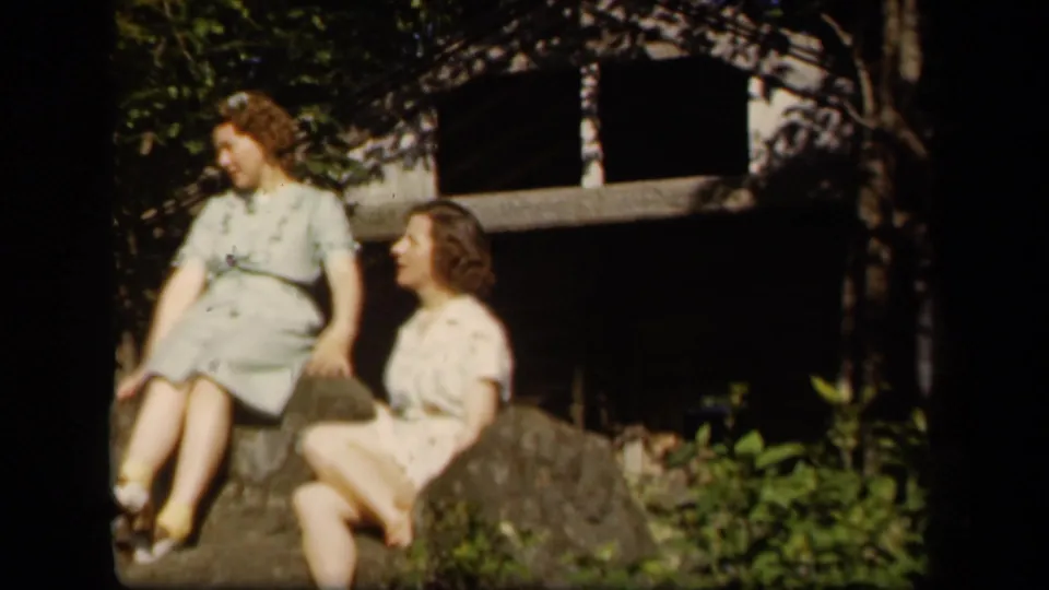 At Skyline Drive, Virginia, two adult women in light dresses appear to converse while seated on rocks in a leafy setting.