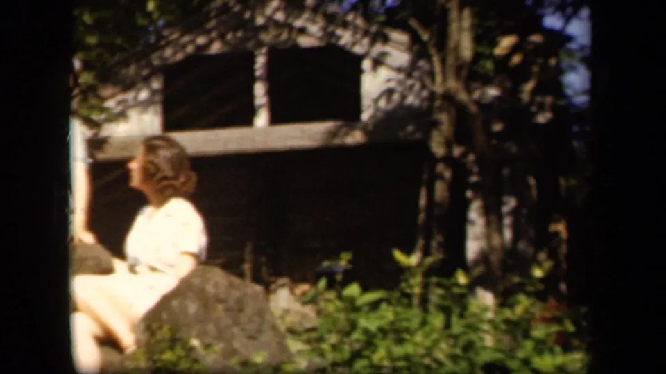 A 1943 Skyline Drive home-movie frame shows two young women conversing near rocks, trees, and a shaded rustic building or shelter.