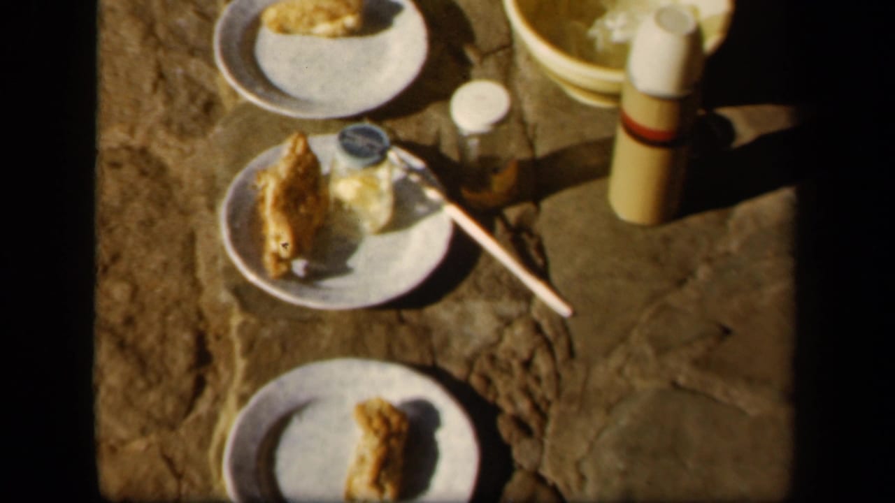 1943 Skyline Drive frame with white plates of food, a thermos, a small jar, a utensil, and a yellow bowl on rough stone.
