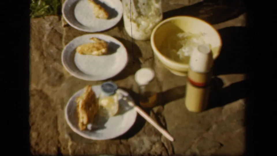 1943 Skyline Drive frame showing the outdoor dinner spread from above, including plates, a yellow bowl, thermos, jar, and utensil.