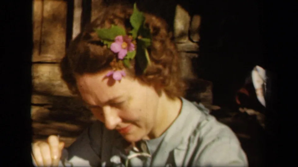 Adult woman with flowers in her hair looks down with a hand raised near the foreground in 1943 Skyline Drive footage.