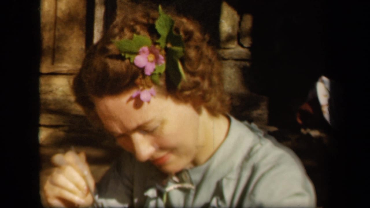 Woman with flowers in her hair looks down while working with her hands on a porch.