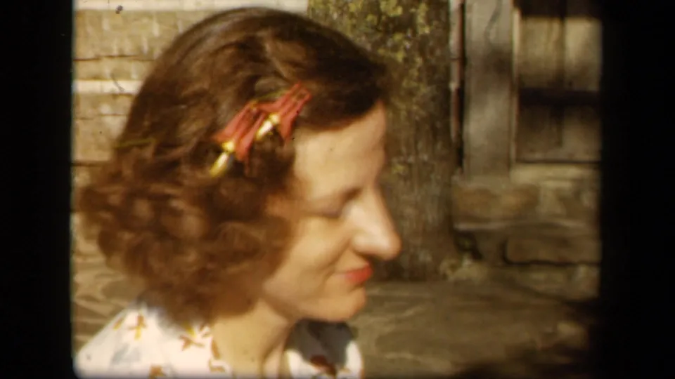In 1943 Skyline Drive home-movie footage, a woman with red floral clips smiles in profile beside stonework and wood siding.