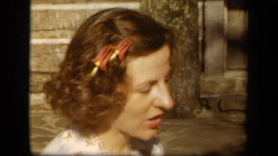 In 1943 Skyline Drive home-movie footage, a woman with red floral hair clips appears to speak while seated outdoors by rustic stone and wood.