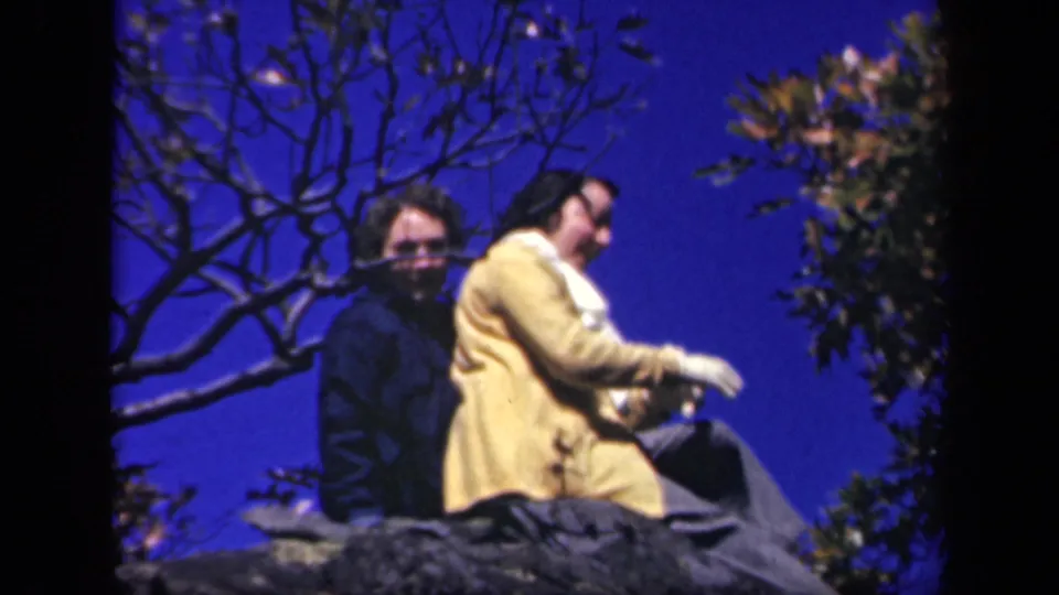 At Skyline Drive, the woman in a yellow coat and white gloves adjusts or removes a scarf while her companion sits behind her.