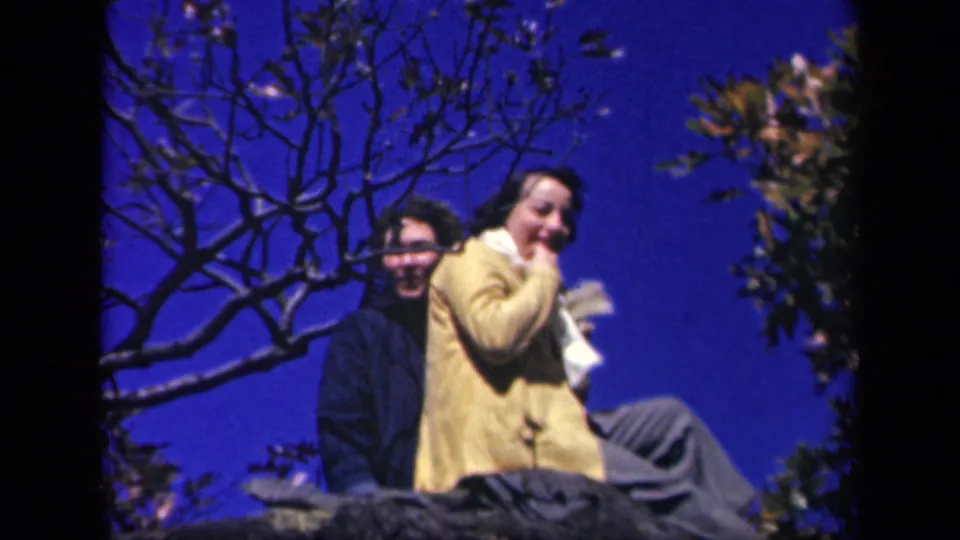 The woman in the yellow coat turns toward the home-movie camera with her scarf near her face as the second woman sits behind her.