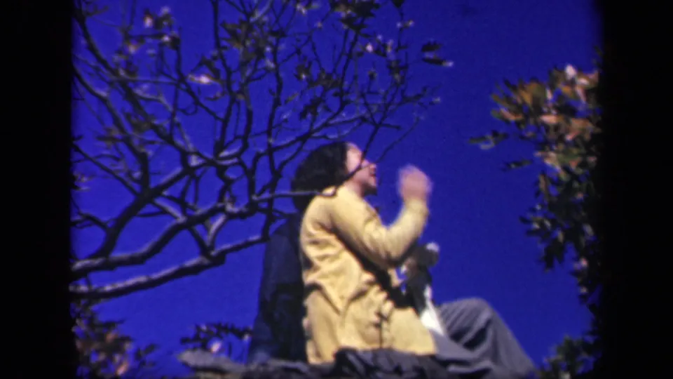 1943 Skyline Drive home-movie frame showing a woman in a yellow sweater seated among branches against a deep blue sky.