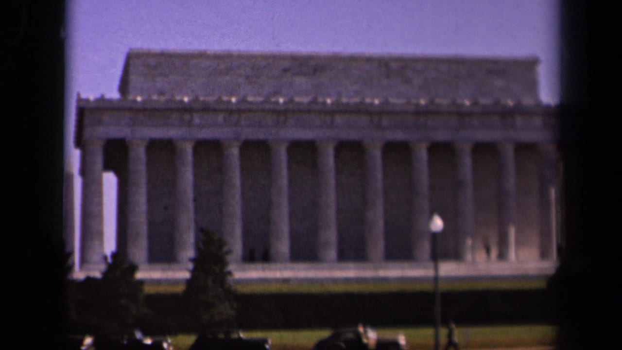 1943 8mm home-movie view of the Lincoln Memorial in Washington, DC, framed with foreground trees, cars, lawn, and pale sky.