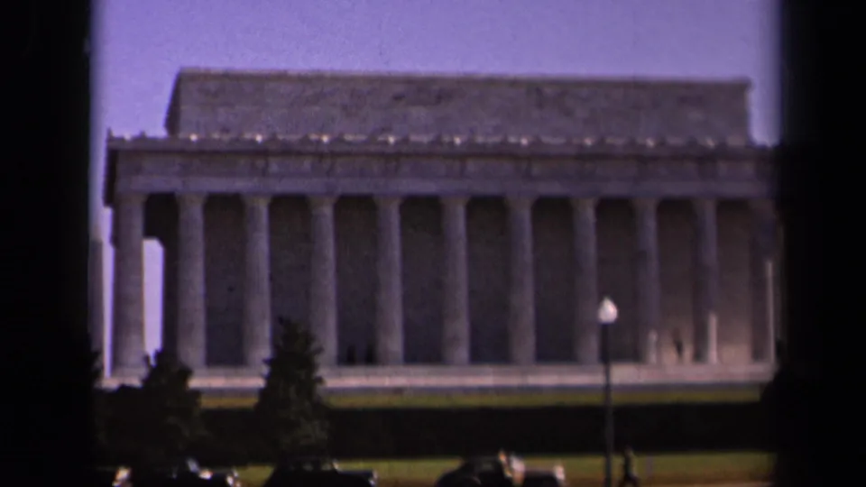 1943 8mm home-movie view of the Lincoln Memorial in Washington, DC, framed with foreground trees, cars, lawn, and pale sky.