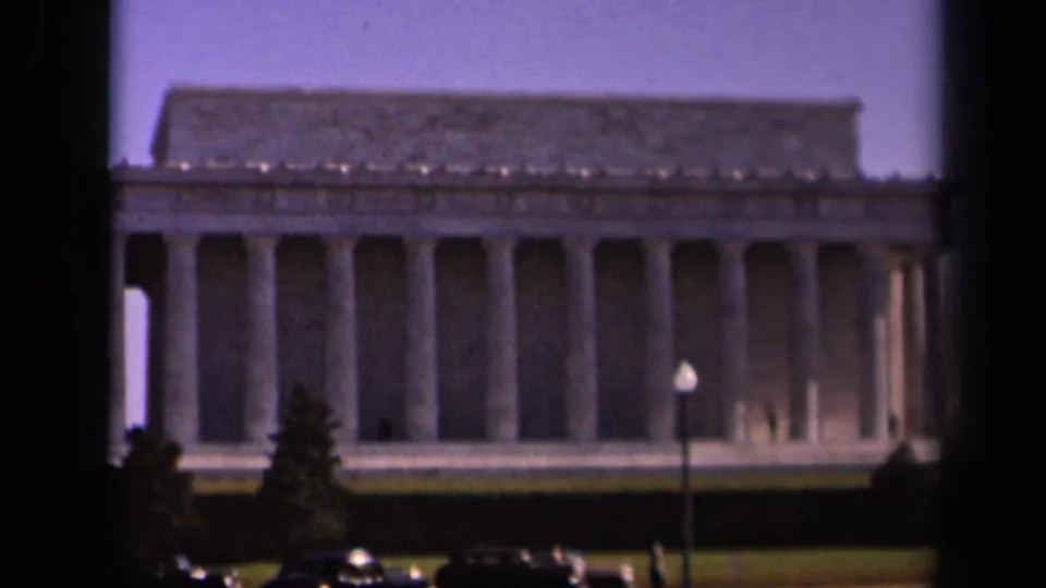 Washington, DC landmark view showing the Lincoln Memorial columns with foreground lawn, trees, lamp post, and classic cars.