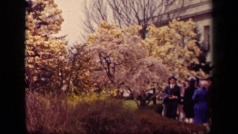 1943 Washington, DC park frame showing visitors in period coats and hats near flowering trees, shrubs, lawn, and a partial building edge.
