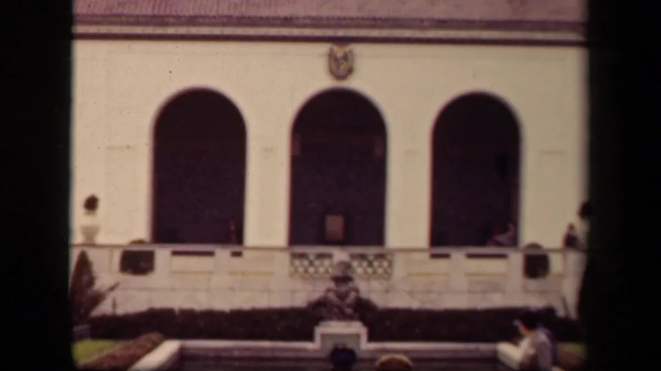 1943 Washington DC home-movie frame showing the Pan-American Union facade with arched openings, balcony rail, emblem, lawn, and distant visitors.