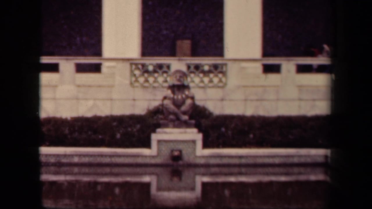 1943 Washington, DC home-movie frame showing a costumed person seated still on a stone pedestal near a formal pool and decorative balustrade.