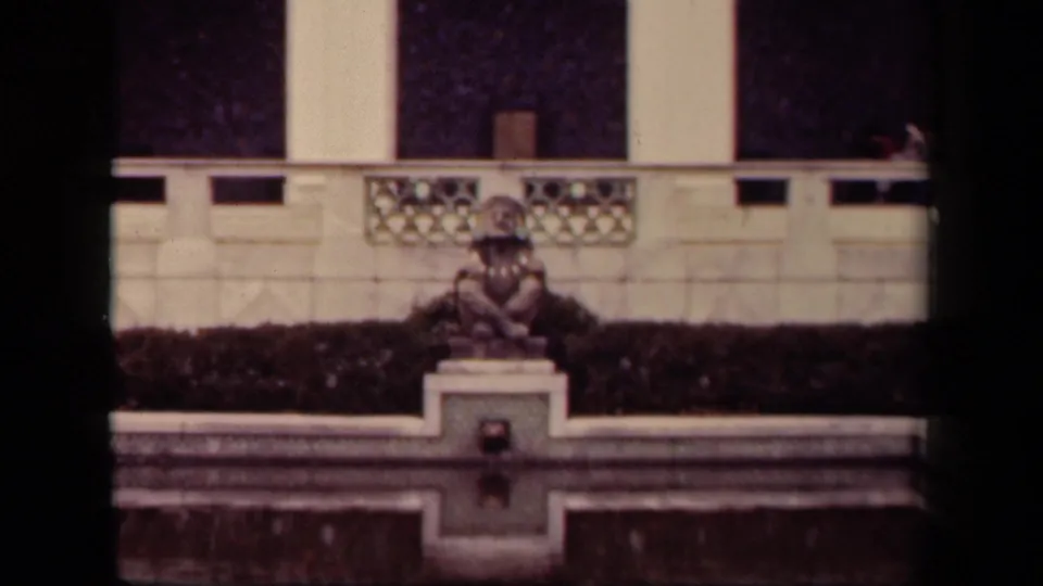1943 Washington, DC home-movie frame showing a costumed person seated still on a stone pedestal near a formal pool and decorative balustrade.