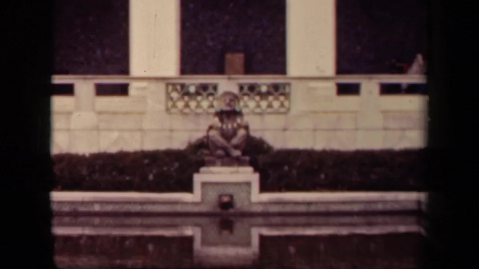 1943 Washington, DC frame with the seated costumed figure centered against a hedge, balustrade, and pale classical architectural backdrop.