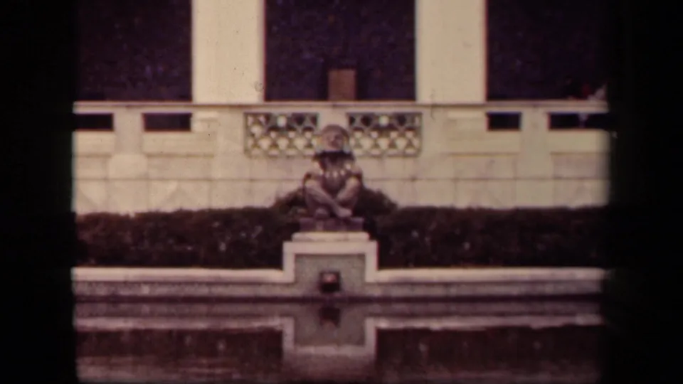 Soft 8mm frame from 1943 Washington, DC showing the same still figure by a reflecting pool with hedge and stone railings behind.