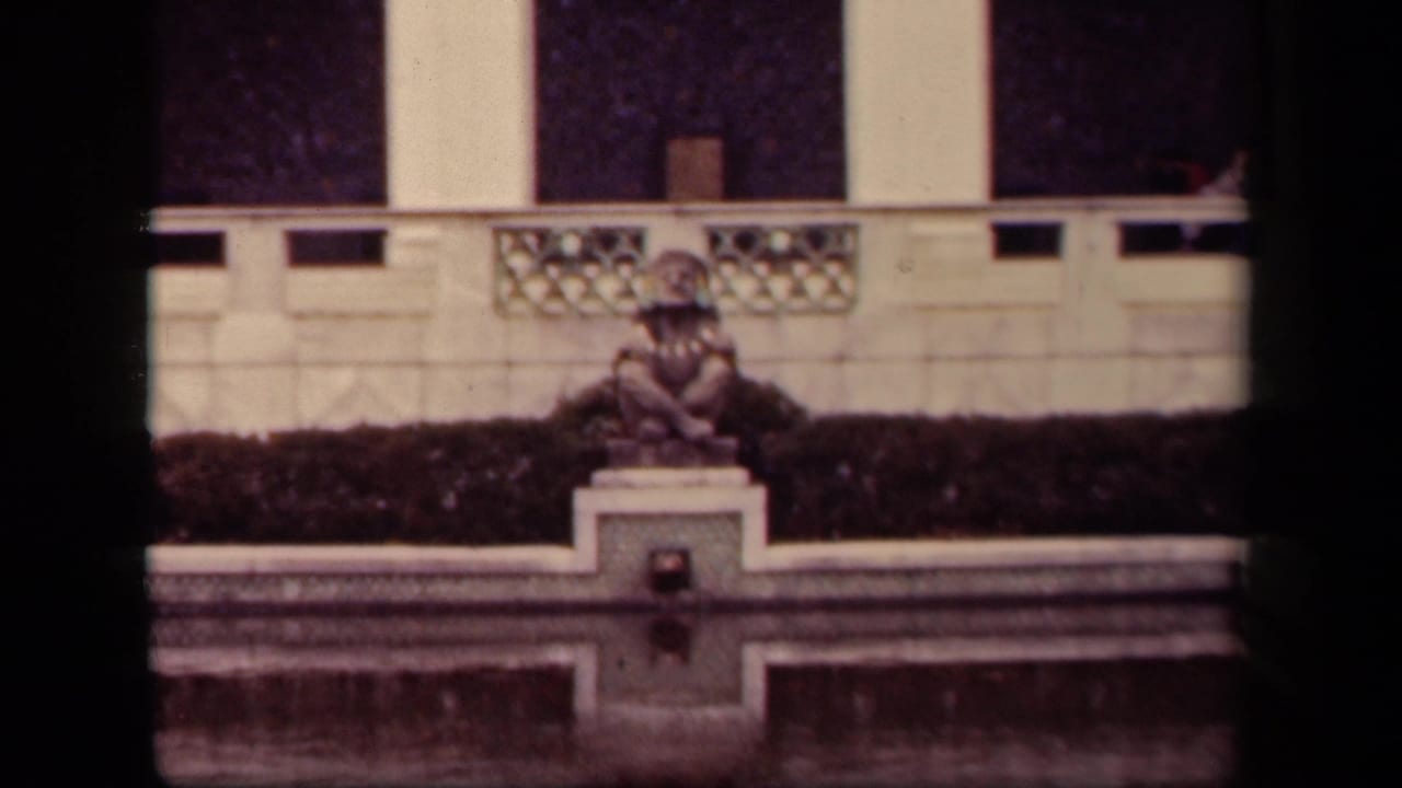 1943 Washington DC home-movie frame showing a small fountain, seated statue, hedge, and pale architectural wall.