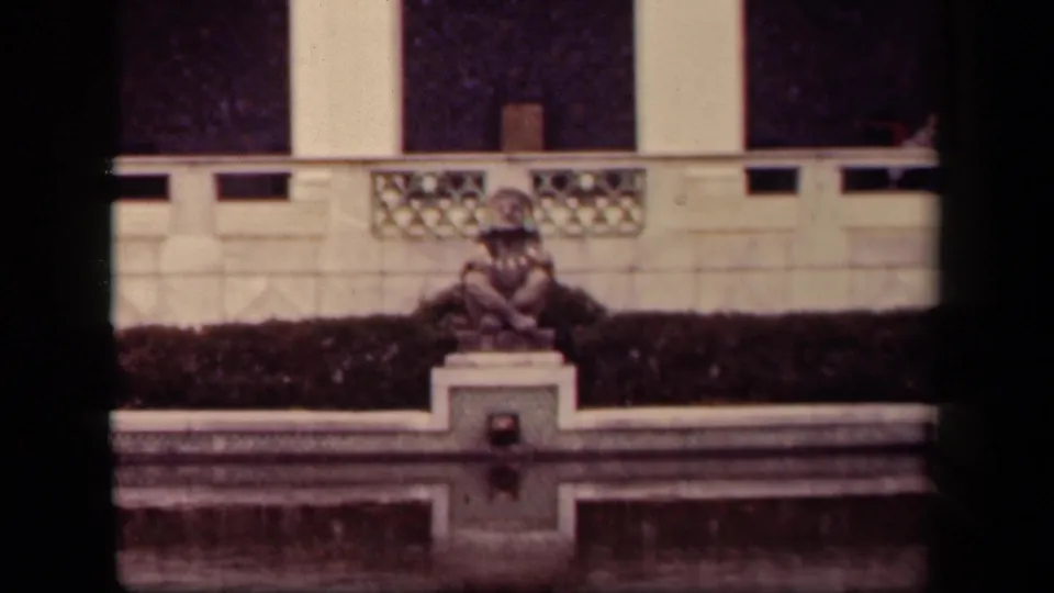 1943 Washington DC home-movie frame showing a small fountain, seated statue, hedge, and pale architectural wall.