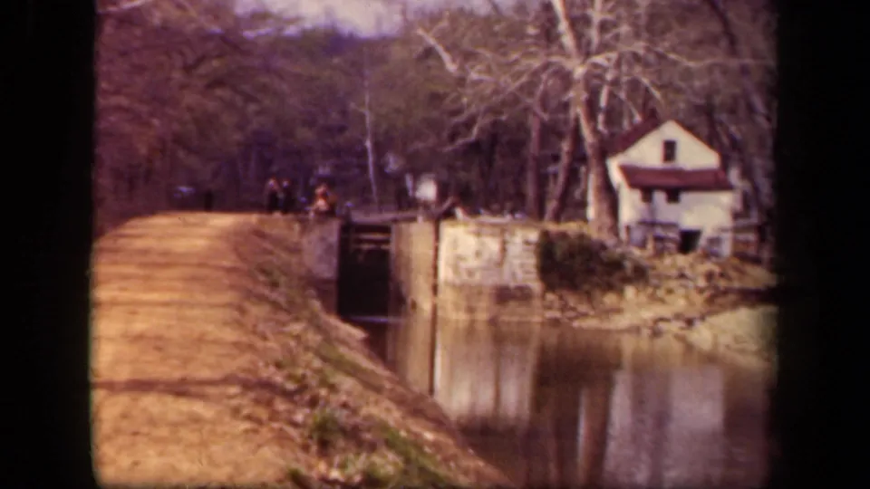 Washington, DC waterway view showing a small white house, lock structure, towpath, bare trees, and distant people in 1943 home-movie footage.