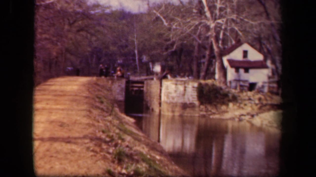 Dirt path beside a canal with stonework, distant walkers, trees, and a small white house.