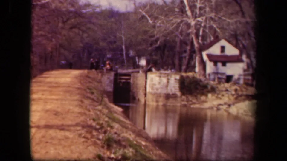 1943 Washington DC 8mm frame showing a canal-side dirt path, stone lock structure, distant pedestrians, bare trees, and a small white house.