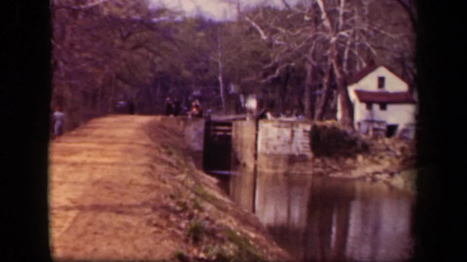 1943 Washington DC home-movie frame of walkers near a stone canal structure, with water, leafless trees, and a small white house nearby.