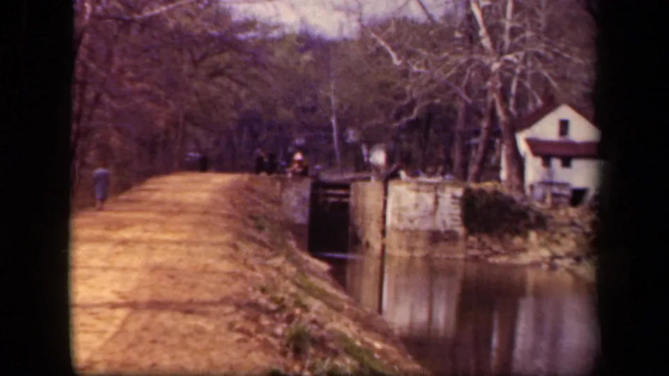 1943 Washington DC 8mm frame showing a person walking along the canal towpath, with stonework, water, trees, and a small white house.