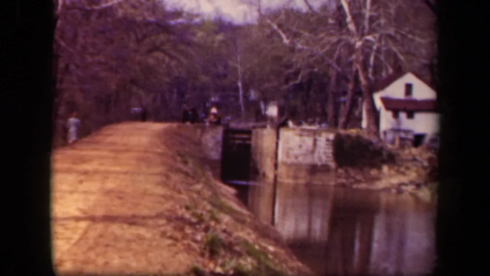 1943 Washington DC home-movie frame of a canal towpath, distant pedestrians, stone lock wall, reflective water, bare trees, and a small white house.