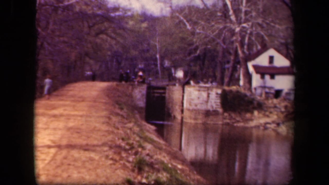 1943 Washington, DC home-movie frame showing a dirt path beside canal-like water, distant pedestrians, bare trees, and a small white building.