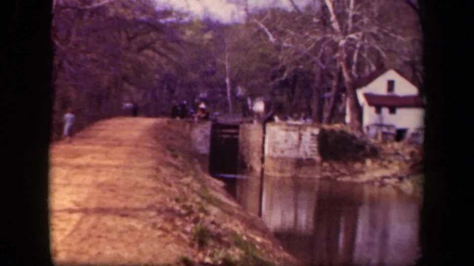 1943 Washington, DC home-movie frame showing a dirt path beside canal-like water, distant pedestrians, bare trees, and a small white building.