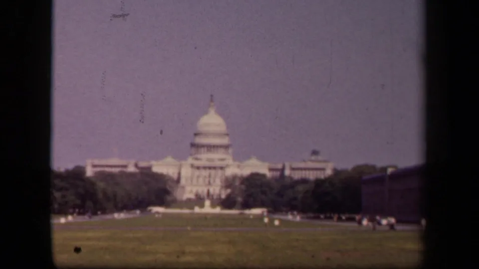 Visitor-shot 8mm view of the U.S. Capitol across the grounds, with trees, lawn, roadway, and small distant people visible.