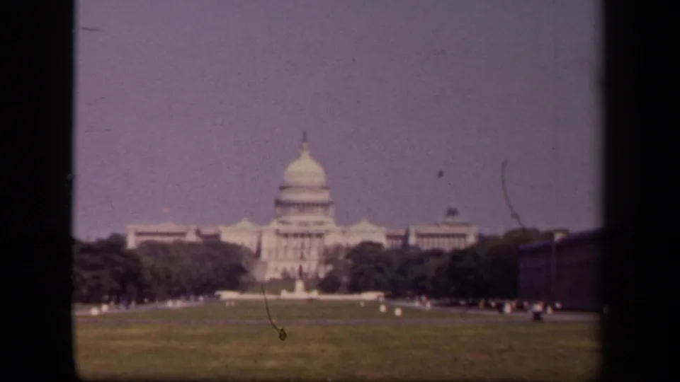 Soft 1940s 8mm shot of the U.S. Capitol from the grounds, showing lawn, trees, distant visitors, and visible film wear.