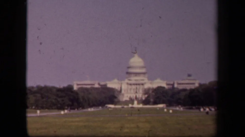 The U.S. Capitol appears from a visitor distance in Washington, DC, with open lawn, trees, and small distant public figures.