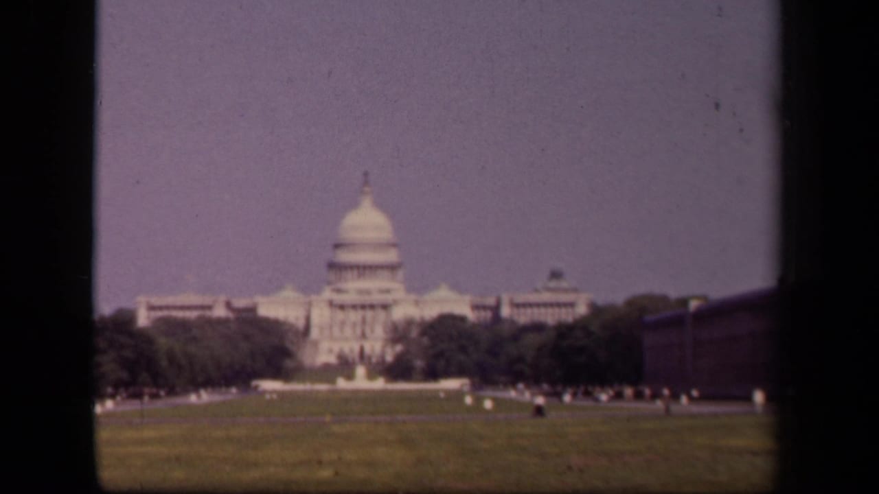 1943 Washington, DC home-movie frame showing the U.S. Capitol across a green lawn, with trees, pale sky, and small distant figures.