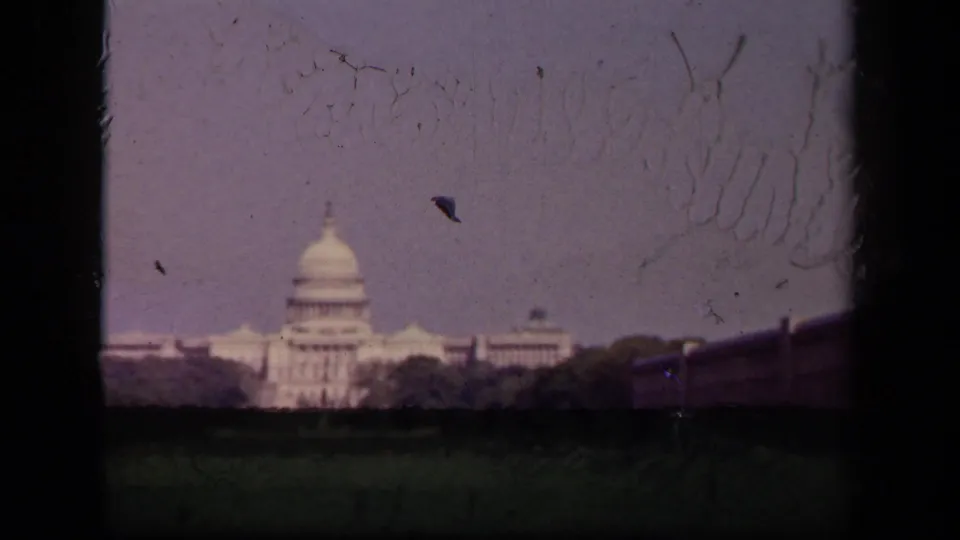 1940s 8mm frame showing the Capitol from the lawn, with visible scratches, dark frame edges, trees, and small distant pedestrians.