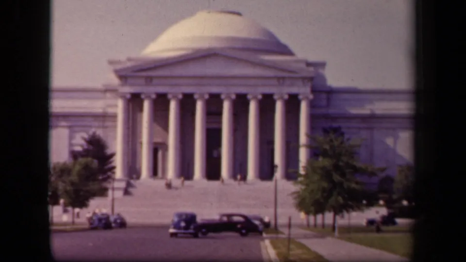 National Art Gallery facade in Washington, DC, with a dark vintage car crossing the foreground and small pedestrians near the steps.