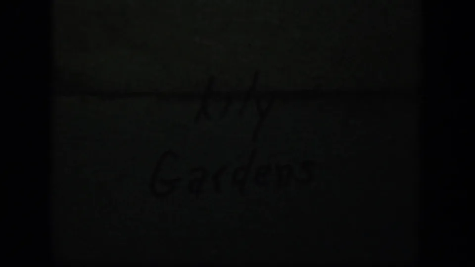 Dim 8mm title-card frame with faint handwriting reading Lily Gardens, partially obscured by dark exposure and film framing.