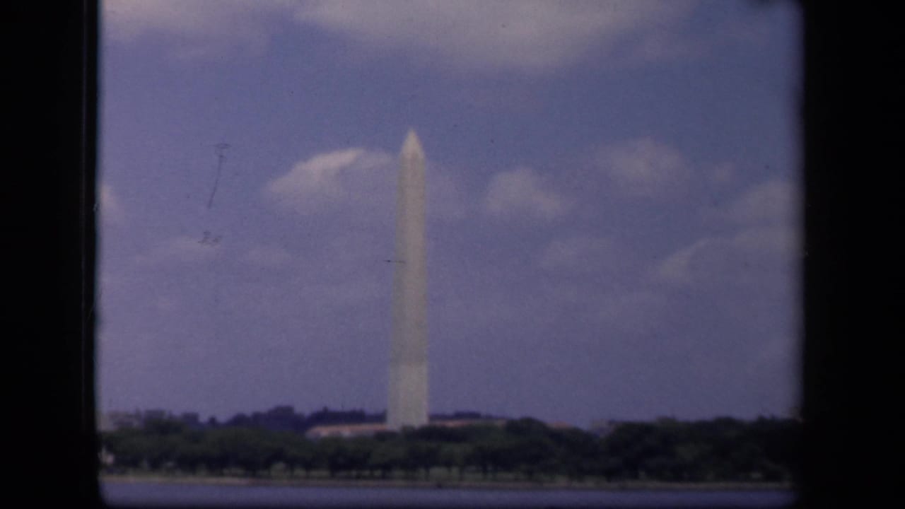 Washington Monument seen beyond water and trees under a cloudy blue sky.