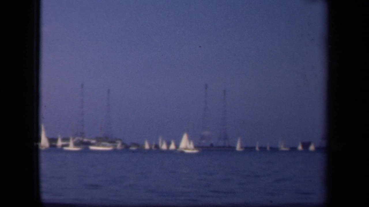 1943 Washington, DC home-movie frame showing small sailboats on blue water with distant utility towers along the horizon.