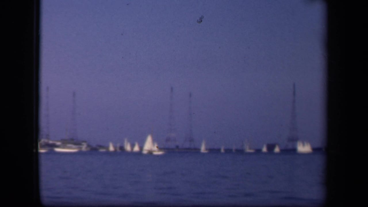 A soft 8mm frame of a sailboat regatta near Washington, DC, with several white sails and tall utility towers in the distance.