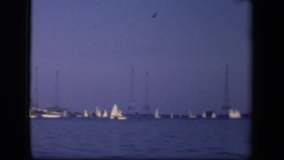 A soft 8mm frame of a sailboat regatta near Washington, DC, with several white sails and tall utility towers in the distance.