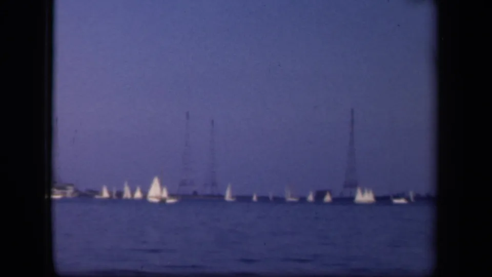 Washington, DC-area 8mm footage frame showing sailboats spread across calm water, with faint power line towers beyond the shoreline.
