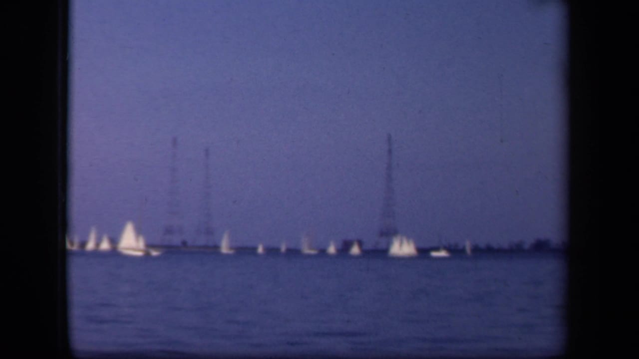 Distant white sailboats on blue water with tall towers on the horizon.